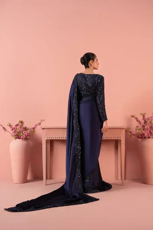 Woman in navy blue embroidered saree with long drape, standing by a table, floral decor