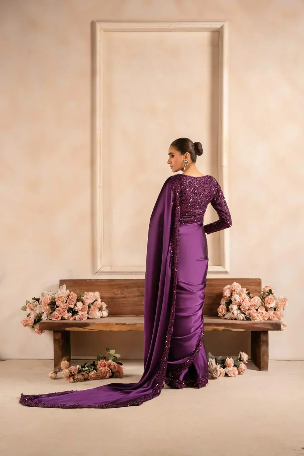 Woman in elegant purple embellished saree standing by a bench with pink roses, fabric fashion