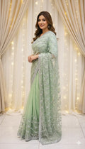 Woman in light green embroidered saree standing indoors with curtain and fairy lights backdrop