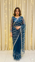 Woman in a blue embroidered saree with sheer sleeves, posing indoors with curtain and string lights