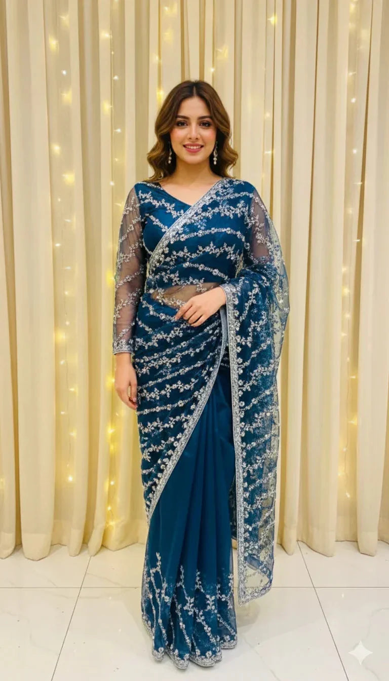Woman in a blue embroidered saree with sheer sleeves, posing indoors with curtain and string lights