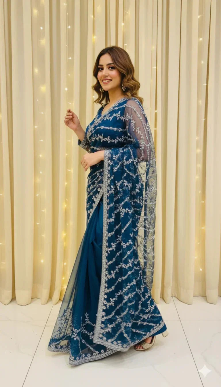 Woman in a blue embroidered saree posing indoors with light curtain backdrop, Sareeium jan fashion