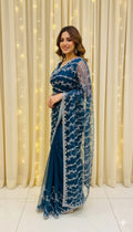Woman in an elegant blue embroidered saree, posing indoors with curtain and fairy lights backdrop