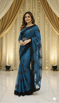 Woman in a blue floral saree with black accents, standing against elegant draped curtains